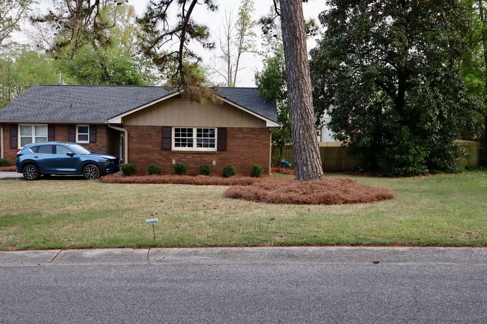 Brick house with car parked in driveway and landscaped yard featuring pine straw.