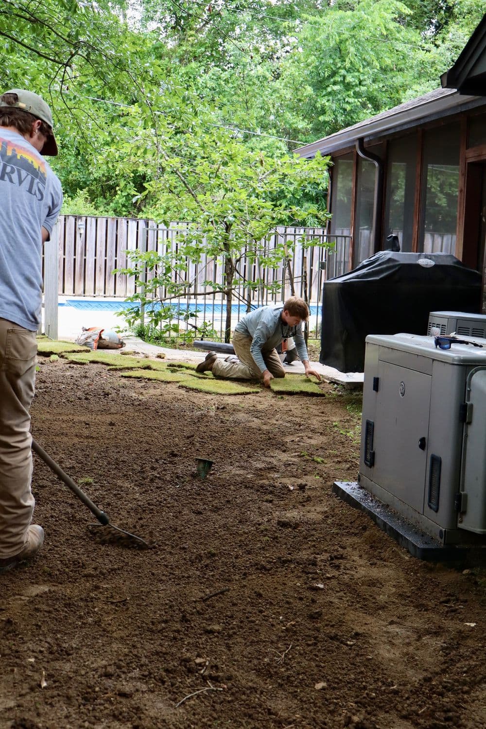 Two workers landscaping a yard, one kneeling on soil and another using a rake. Green trees in background.