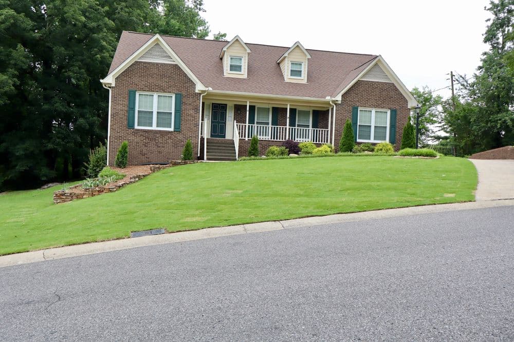 Brick house with a landscaped yard and porch on a sloped driveway in a suburban neighborhood.